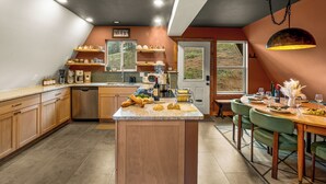 Een keuken met houten kasten, een centraal eiland met etenswaren, een eetgedeelte met tafel en stoelen, en uitzicht door een raam.