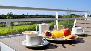 Un desayuno preparado en una mesa al aire libre con una vista panorámica, que incluye una taza blanca, un vaso de zumo de naranja, una manzana roja y un plato con bollería.