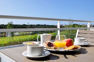 Un desayuno preparado en una mesa al aire libre con una vista panorámica, que incluye una taza blanca, un vaso de zumo de naranja, una manzana roja y un plato con bollería.