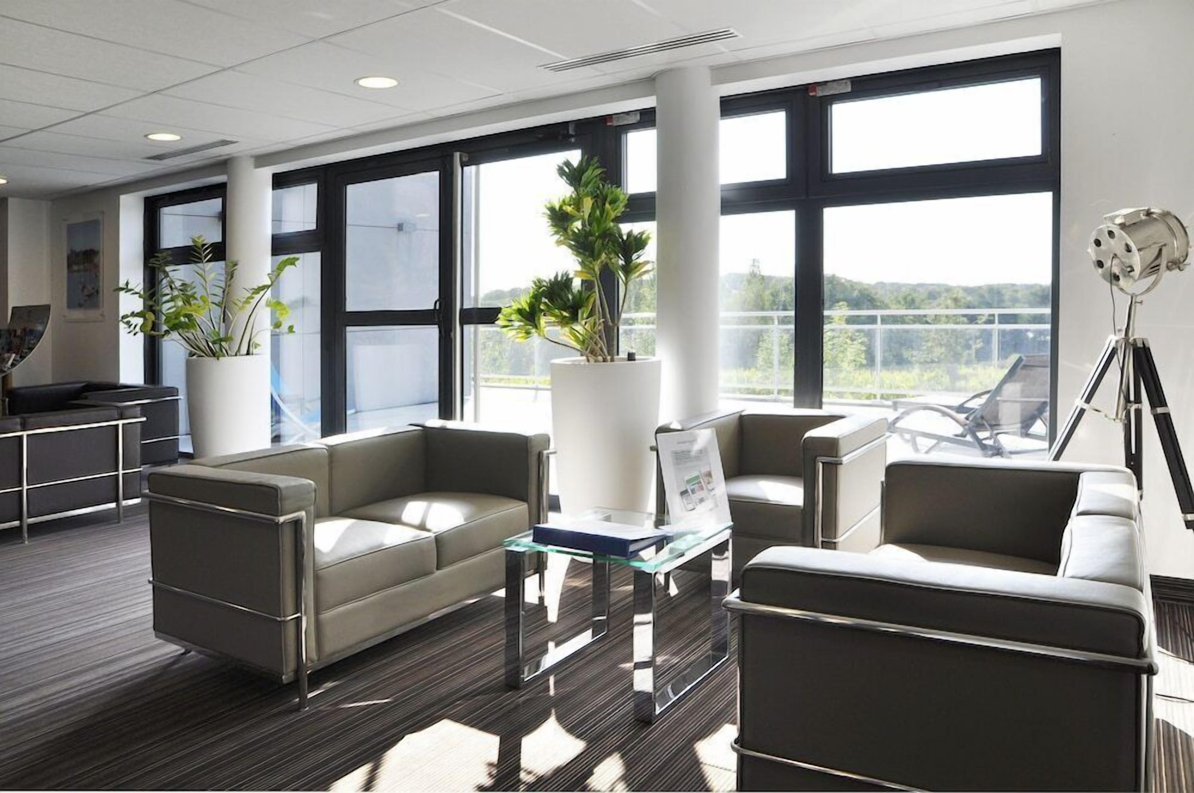 A modern office space with two sofas, a glass table, potted plants, and large windows.