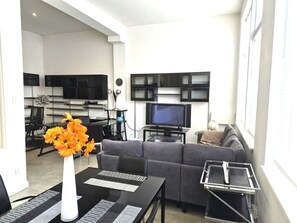 Ein modernes Wohnzimmer mit einem grauen Sofa, einem Esstisch mit Blumenvase, einem flachbildfernseher, der über einer schwarzen Unterhaltungsanlage angebracht ist, und einer Küche im Hintergrund.