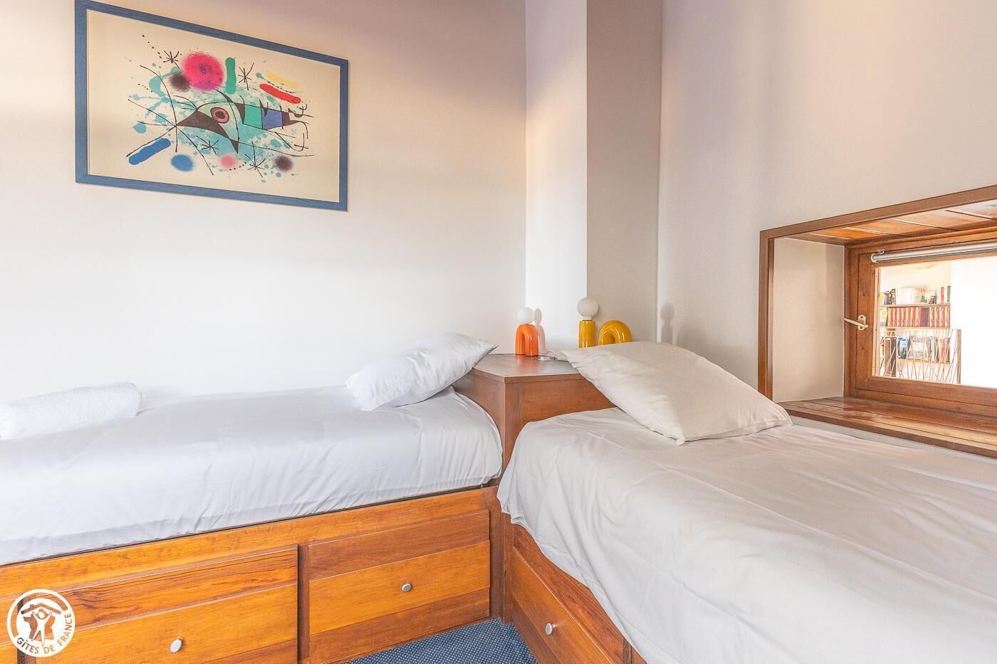 A room with two beds, wooden headboards, a framed abstract painting, a nightstand with orange and yellow items, and a window with a view of bookshelves.