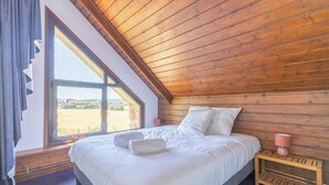 Ein Schlafzimmer mit Holzbalkendecke, einem großen Fenster, einem Bett mit weißer Bettwäsche und einem Nachttisch mit Lampe.