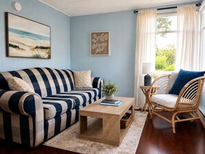 A living room with a striped sofa, a wooden coffee table, a wicker chair, and a window with curtains. - Little Tracadie Retreat (Grand Tracadie)