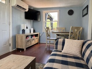 A living room with a television, wooden furniture, a dining table with chairs, and a window with a view. - Little Tracadie Retreat (Grand Tracadie)