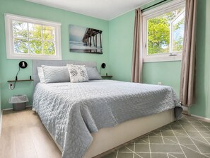 A bedroom with a bed, two bedside tables, a wall-mounted lamp, a picture, and a window with curtains. - Little Tracadie Retreat (Grand Tracadie)