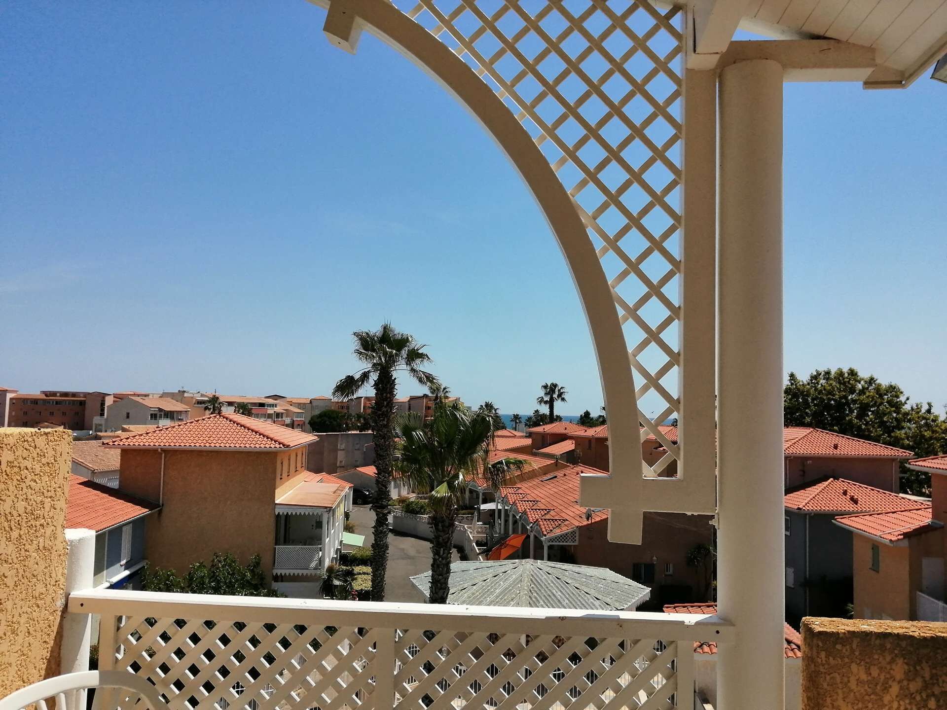 A balcony with a lattice structure, overlooking a residential area with red-tiled roofs and palm trees.