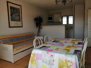 A room with a sofa, a dining table with a floral tablecloth, a potted plant, a microwave, and a door. - Family 2 bed apartment / Rochelongue (CAP D AGDE)
