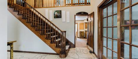A spacious hallway with a wooden staircase, stone flooring, and a large glass door.