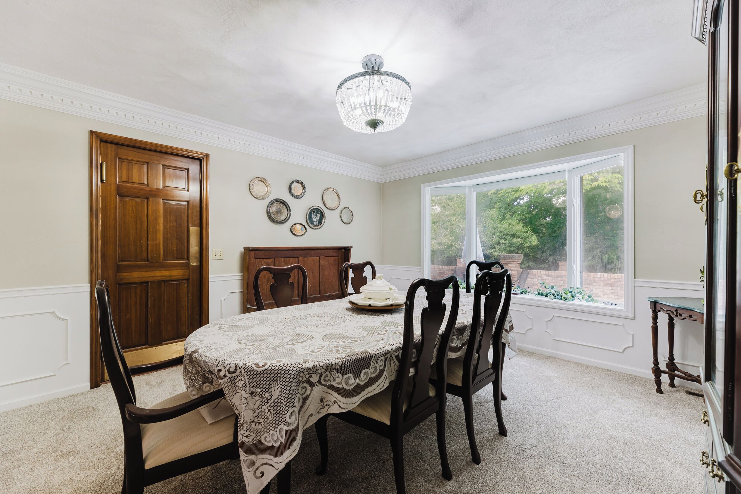 A dining room with a chandelier, a wooden door, and a window with a view of greenery.