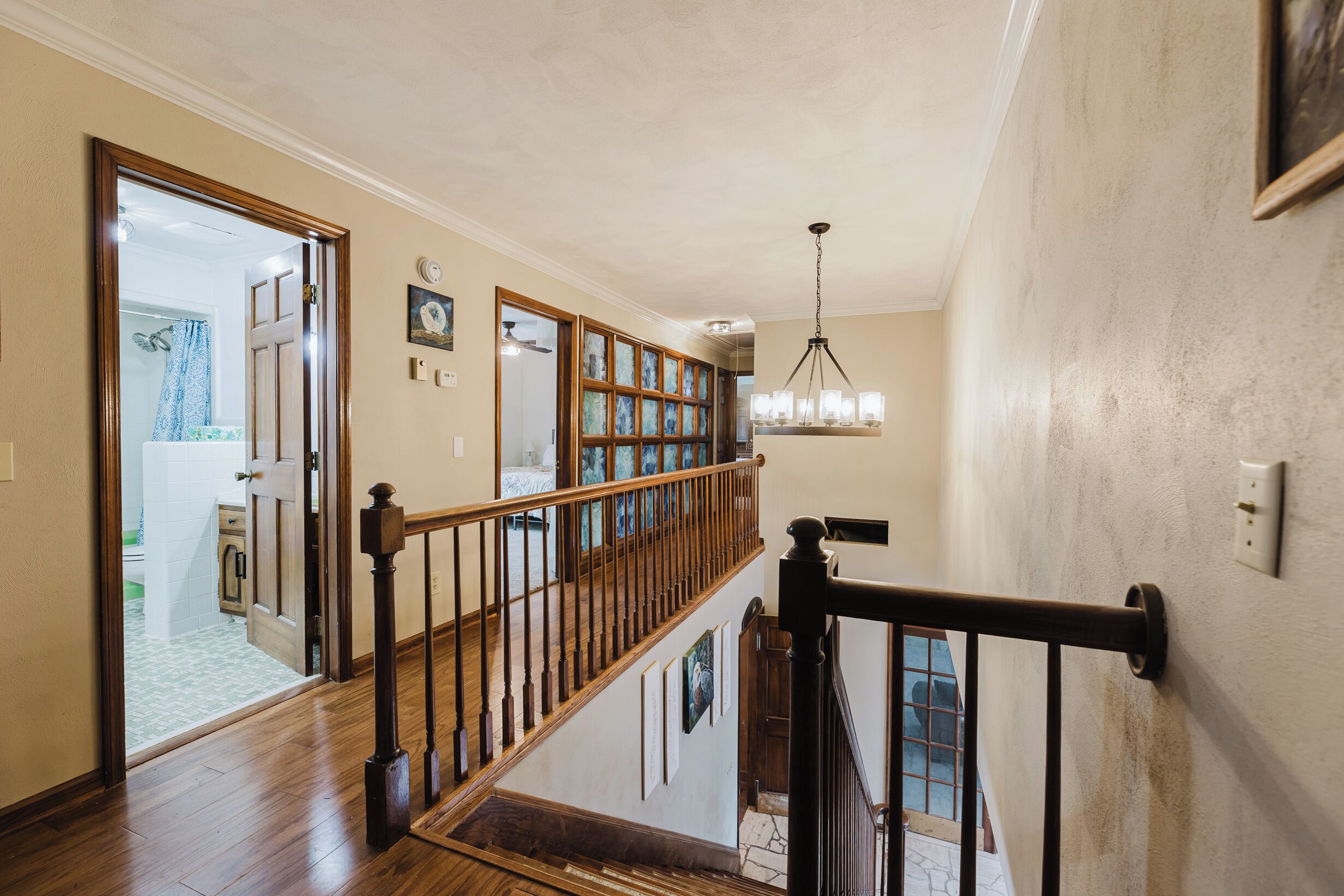 A staircase with wooden railings and a chandelier, leading to a room with a bed and a bathroom visible through an open door.