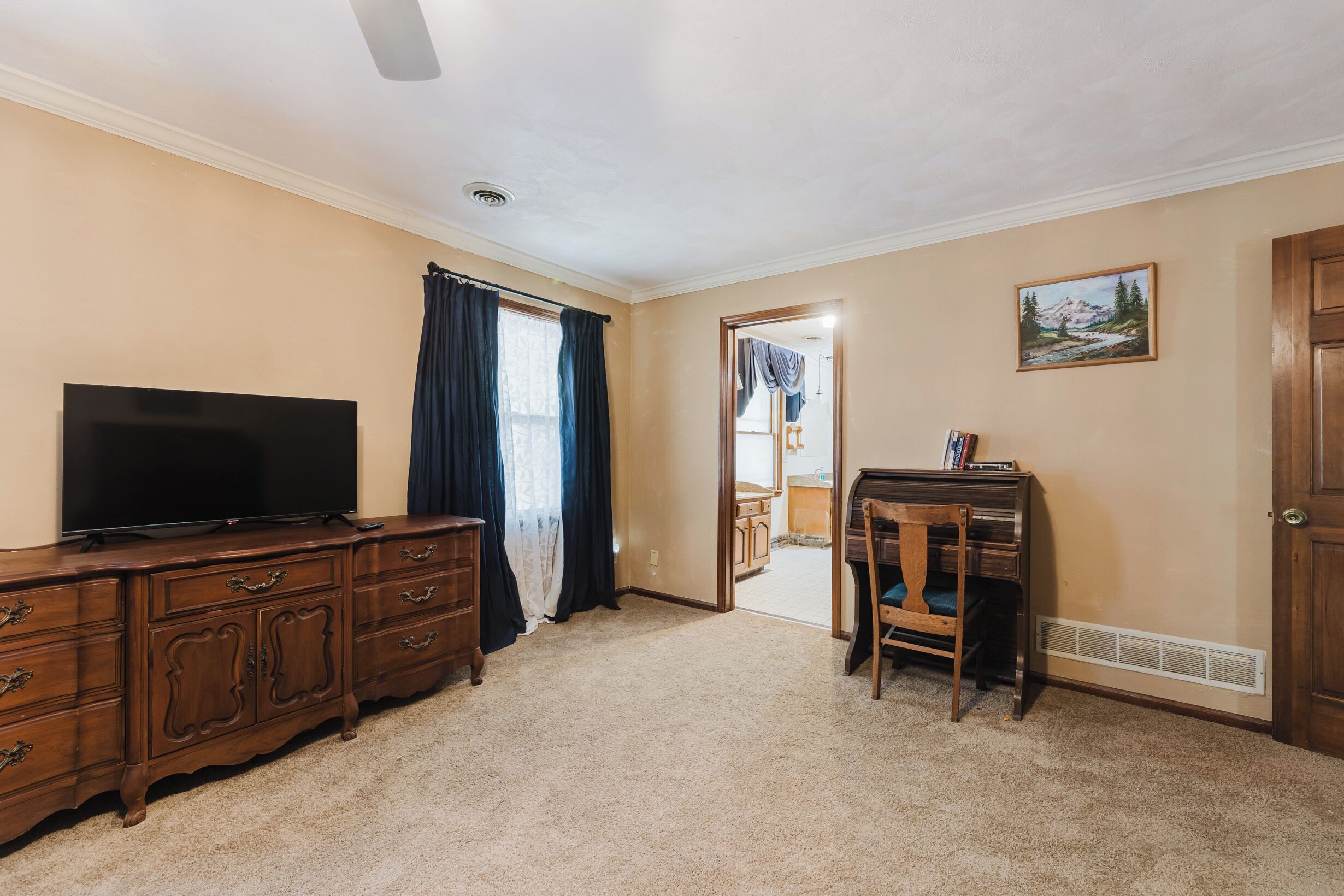 A room with a flat-screen TV on a wooden dresser, a chair, a desk with a computer, a painting on the wall, and a door leading to another room.
