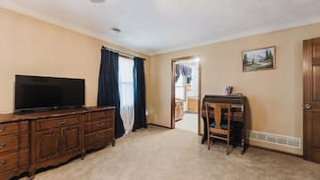 A room with a flat-screen TV on a wooden dresser, a chair, a desk with a computer, a painting on the wall, and a door leading to another room.