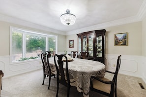 Une salle à manger avec une table ronde, des chaises, un lustre, un buffet et une fenêtre donnant sur un paysage verdoyant.