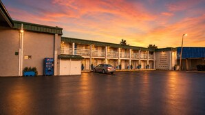 Um motel com um carro estacionado no estacionamento, uma máquina de refrigerantes da Pepsi e uma placa de boas-vindas a veteranos, viajantes, trabalhadores, famílias e todas as culturas.