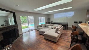 Een moderne woonkamer met een modulaire bank, een open haard, een grote schuifpui naar een achtertuin, en een keuken met een bar en barkrukken.