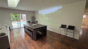 Een ruime kamer met een houten eettafel, twee barkrukken, een bank en een groot raam met uitzicht op een zwembad en groen.