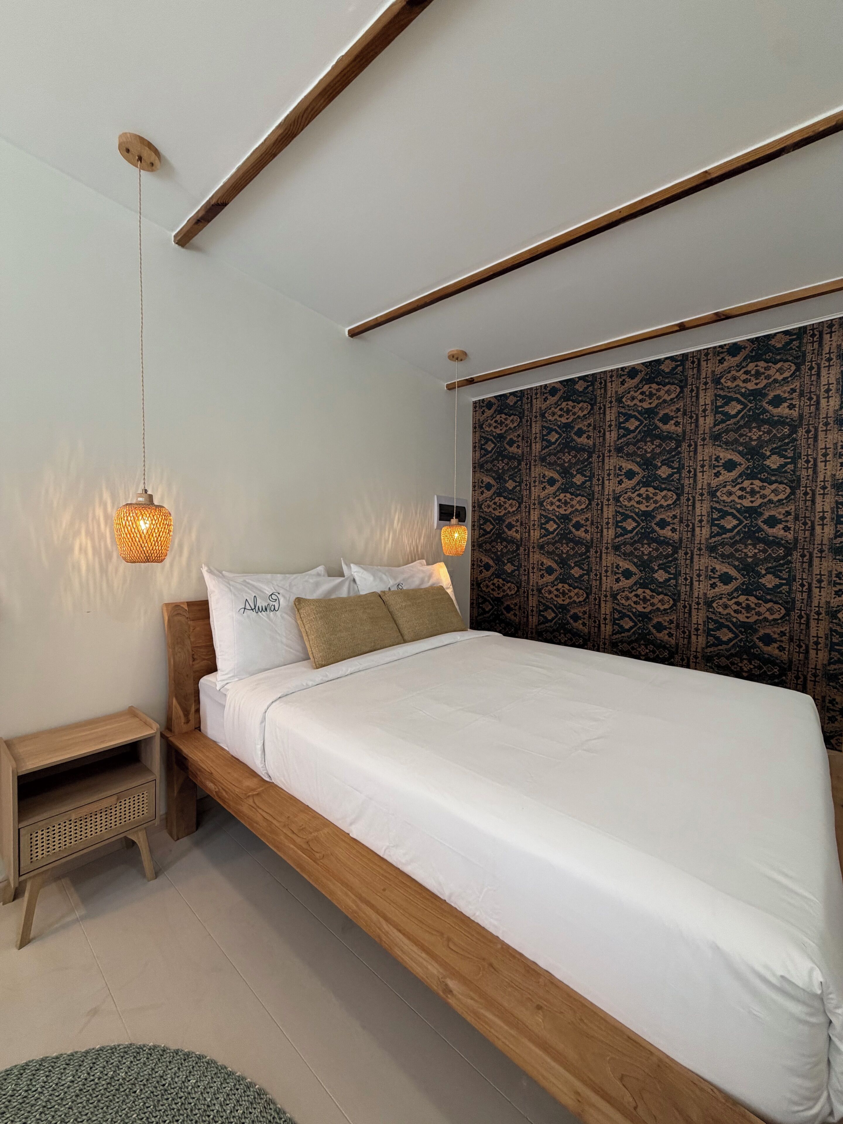 A bedroom with a wooden bed frame, white bedding, a patterned headboard, and two hanging lamps.