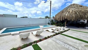 A swimming pool with lounge chairs, a thatched-roof structure, and a white wall. - Boutique Studio | King Bed & Pool | Unit 1 | Aluna (Savaneta)
