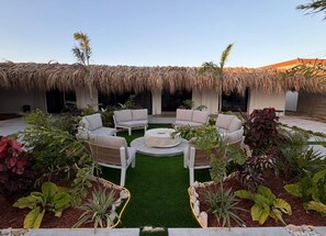 A circular seating area with a fire pit, surrounded by lush greenery and a thatched roof structure. - Boutique Studio | King Bed & Pool | Unit 1 | Aluna (Savaneta)