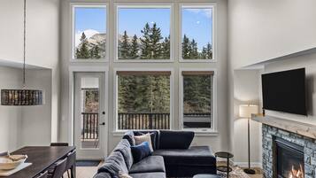 Un salon moderne avec une cheminée, un grand téléviseur et une vue sur les arbres grâce à de grandes fenêtres.