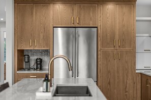 A modern kitchen with wooden cabinets, a stainless steel refrigerator, a coffee maker, and a marble countertop. - Walk to Town | Luxury Home with Private Hot Tub (Canmore)