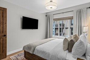Un dormitorio con cama, televisión montada en la pared, ventana con cortinas y puerta de madera.