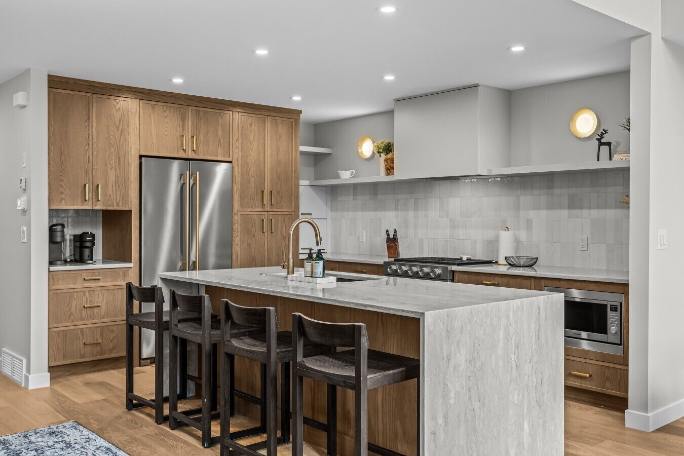 A modern kitchen with wooden cabinets, a stainless steel refrigerator, a central island with bar stools, a gas stove, and a built-in oven.