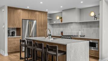 A modern kitchen with wooden cabinets, a stainless steel refrigerator, a central island with bar stools, a gas stove, and a built-in oven.