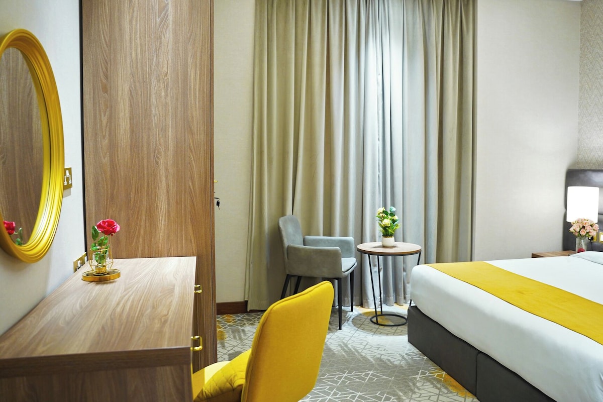 A hotel room with a bed, a desk with a mirror, a chair, a small table with flowers, and a nightstand with a lamp.