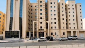 Un hotel de varios pisos con entrada central, fachada de vidrio moderna y un estacionamiento con varios autos.
