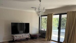 A spacious room with a large flat-screen TV, a chandelier, and a sliding glass door leading to a balcony. - Luxury Fully Furnished Acreage Retreat – Dural (Dural)