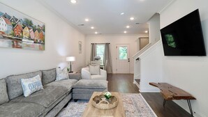 Ein Wohnzimmer mit grauem Sofa, einem Holztisch, einem an der Wand befestigten Fernseher und einer Treppe zu einem anderen Raum.