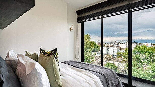 Un dormitorio con un ventanal grande, una cama con muchas almohadas y vistas a un paisaje urbano.