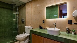 Et badeværelse med glasbrusekabine, toilet, vask med spejl over og en grøn flisegulv.