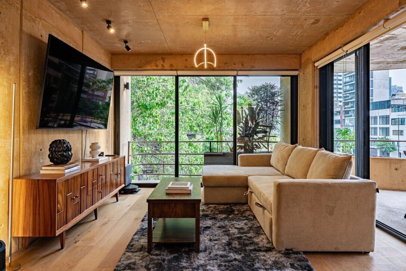 Ein modernes Wohnzimmer mit Holzboden, einem Flachbildfernseher, einem beige farbenen Sofa, einer Holzkommode und großen Fenstern mit Blick auf Grünflächen und Gebäude.