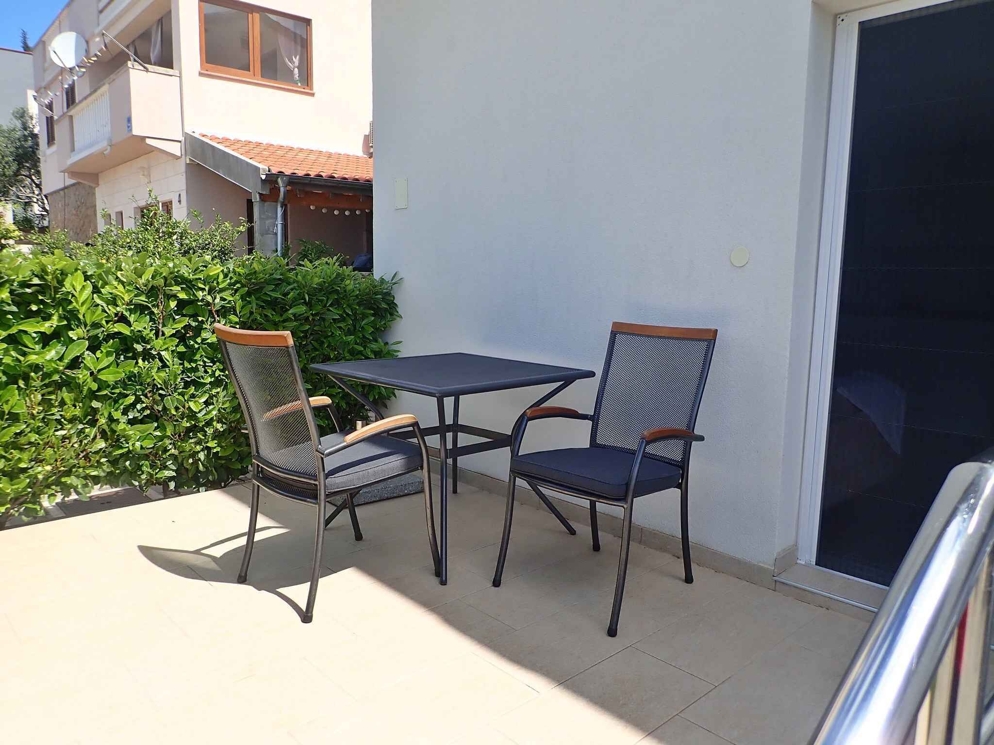 An outdoor patio with a table and two chairs, a white wall, and a green hedge.