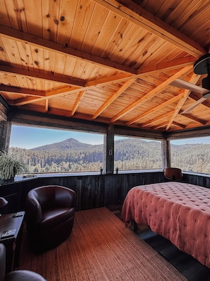 Ein Holzblockhaus-Interieur mit einem Bett, einem Sessel und Blick auf einen bewaldeten Berg.
