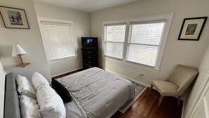 Une chambre à coucher avec un lit, un téléviseur, une chaise et deux fenêtres équipées de stores.