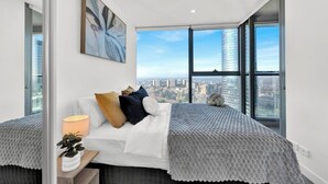 Un dormitorio moderno con un ventanal grande, una cama con un edredón texturizado, una mesita de noche con una lámpara y una planta en maceta.