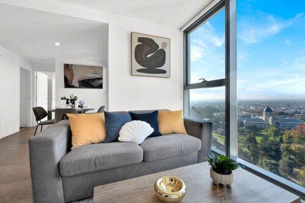 A modern living room with a grey sofa, a large window with a city view, and a dining area in the background. - Incredible Views - Bright & Spacious CBD Escape (Melbourne)