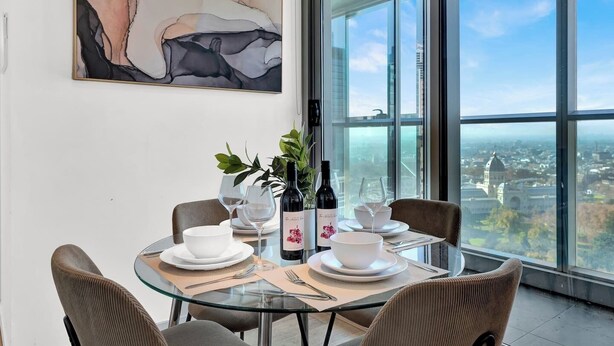 Un comedor con una mesa de vidrio, platos blancos y botellas de vino, con vista a un paisaje urbano.