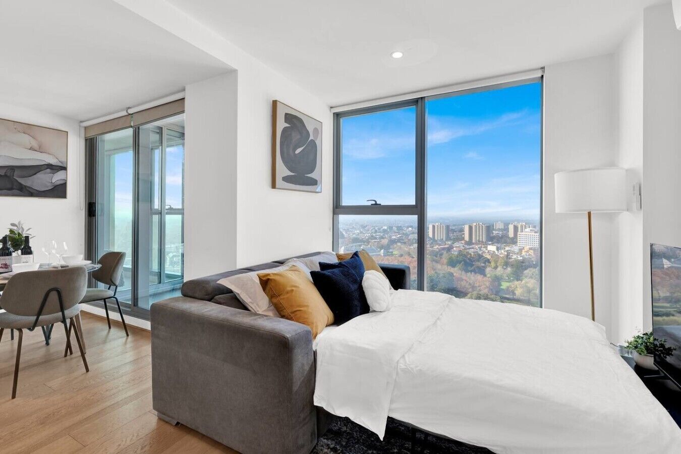 A modern bedroom with a large bed, a grey sofa, a dining area with chairs, and a city view through large windows.