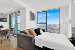 A modern bedroom with a large bed, a grey sofa, a dining area with chairs, and a city view through large windows. - Incredible Views - Bright & Spacious CBD Escape (Melbourne)