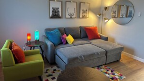 A modern living room with a grey sofa, colorful cushions, a green armchair, a small side table with a lamp, and framed animal prints on the wall.