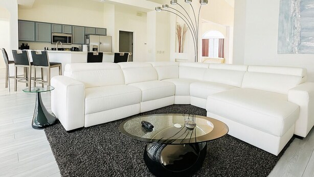 A modern living room with a white sectional sofa, a glass coffee table, and a kitchen in the background. - Pool Oasis Near Disney, Games & TV, Family Time (Davenport)