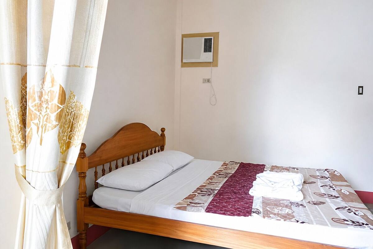 A single bed with a wooden headboard, a white pillow, a patterned bedspread, and a wall-mounted air conditioning unit.