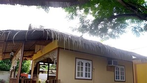 A single-story building with a thatched roof, a front porch, and a small garden area. - La Petite Pension (Puerto Princesa)