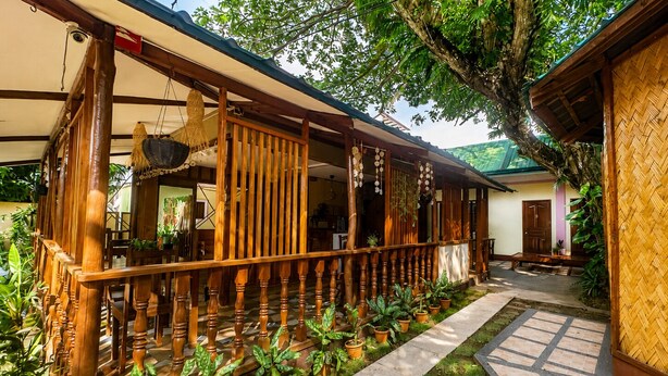 A covered wooden porch with a railing, potted plants, and a pathway leading to a door. - La Petite Pension (Puerto Princesa)