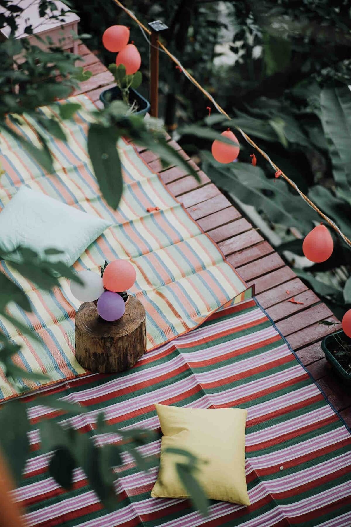 Un espace extérieur accueillant avec des coussins à rayures, un tronc d’arbre servant de siège et des ballons rouges.